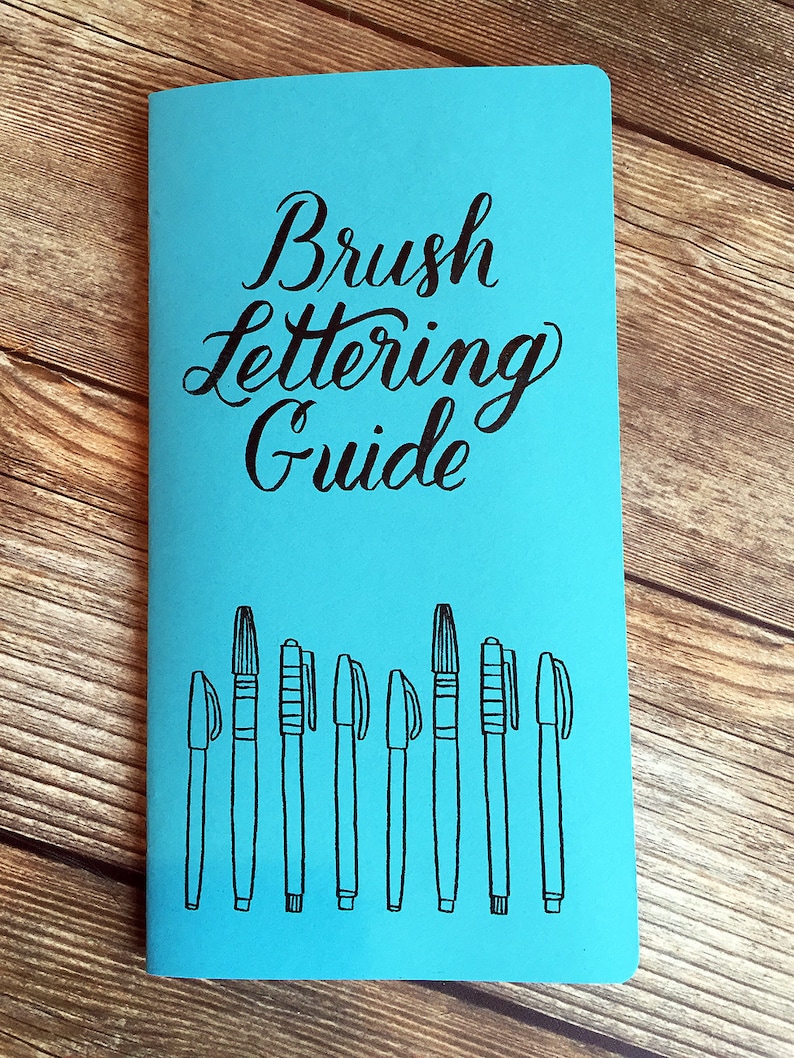May include: A blue notebook with the text "Brush Lettering Guide" in black cursive lettering. The notebook has a row of ten black brush pens drawn on the front.