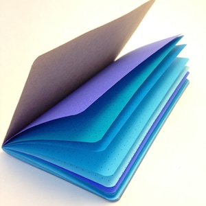 May include: A notebook with a purple cover and blue pages. The pages are layered in shades of blue, creating a gradient effect. The notebook is open and the pages are fanned out.
