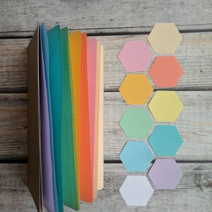 Puede incluir: Un cuaderno marr&oacute;n con p&aacute;ginas de colores del arco&iacute;ris y un patr&oacute;n de puntos. Junto al cuaderno hay varios recortes de papel con forma de hex&aacute;gono de colores en tonos rosa, naranja, amarillo, verde, azul y morado.