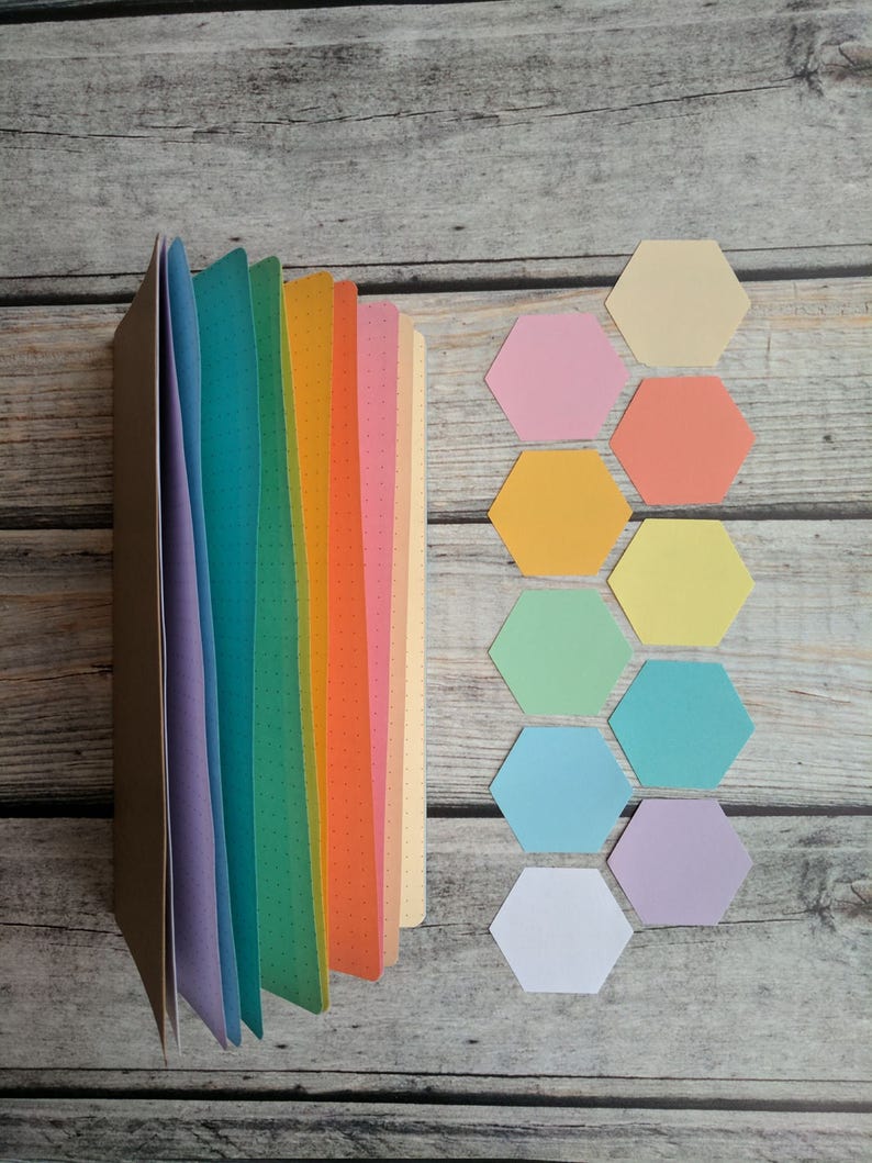 Puede incluir: Un cuaderno marr&oacute;n con p&aacute;ginas de colores del arco&iacute;ris y un patr&oacute;n de puntos. Junto al cuaderno hay varios recortes de papel con forma de hex&aacute;gono de colores en tonos rosa, naranja, amarillo, verde, azul y morado.