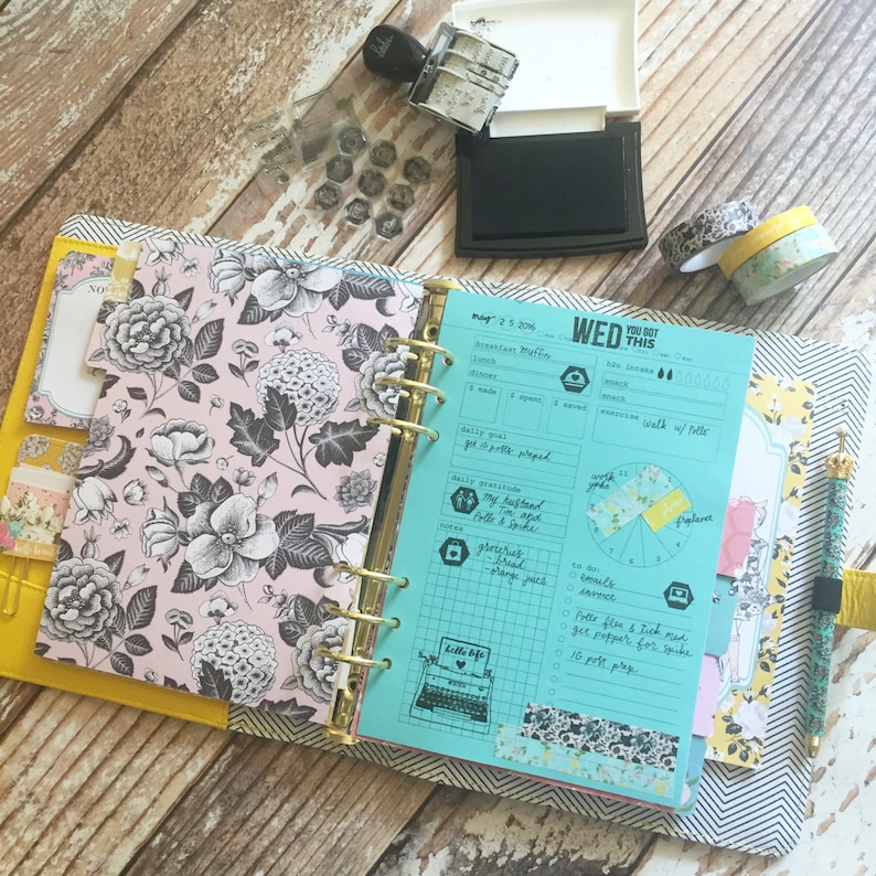 以下が含まれることがあります： ターコイズブルーのプランナーで、表紙には花柄のデザインと「WED YOU GOT THIS」の文字が書かれています。プランナーは、週ごとのスプレッドに開かれており、毎日の感謝のセクション、やることリスト、進捗状況を追跡するための円グラフがあります。