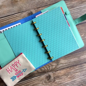 May include: A teal blue planner with gold rings and a turquoise cover. The planner is open to a page with a grid pattern. A small beige pouch with the text "HAPPY is this way" is in the foreground.