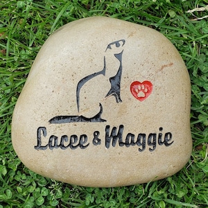 May include: A light brown stone with a black silhouette of a ferret and a red paw print heart. The stone is engraved with the text "Lace & Maggie".