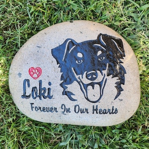 May include: A light brown stone with a black silhouette of a dog's head and the words "Loki Forever In Our Hearts" and a red paw print heart.