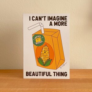 May include: A rectangular art print featuring a cartoon juice box with a smiling corn cob. The juice box has a straw and the text "I CAN'T IMAGINE A MORE BEAUTIFUL THING". A sticker on the box reads "now with butter".