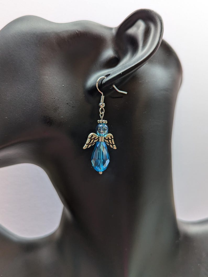 Puede incluir: Pendientes de aro de plata con alas de &aacute;ngel y un cristal azul en forma de l&aacute;grima.