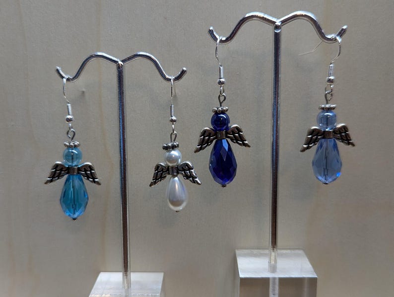 Puede incluir: Tres pares de pendientes colgantes con forma de &aacute;ngel. Un par presenta un cristal azul, otro una perla blanca y el tercero un cristal azul oscuro.