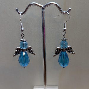 Puede incluir: Un par de pendientes colgantes con un dije de &aacute;ngel plateado y un cristal azul en forma de l&aacute;grima.