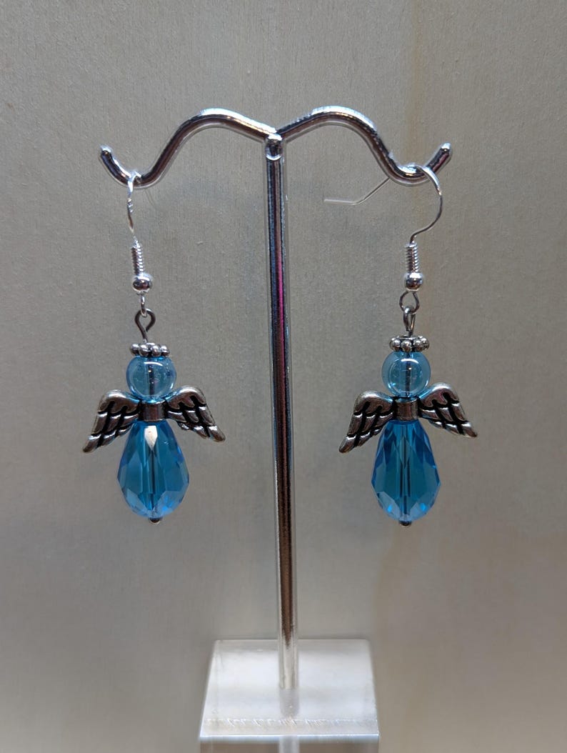 Puede incluir: Un par de pendientes colgantes con un dije de &aacute;ngel plateado y un cristal azul en forma de l&aacute;grima.