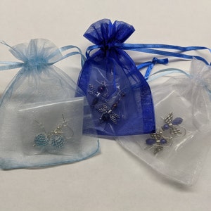 Puede incluir: Tres peque&ntilde;as bolsas de organza con cintas de diferentes colores. Cada bolsa contiene un par de pendientes. Una bolsa es azul, otra es blanca y la &uacute;ltima es azul claro.