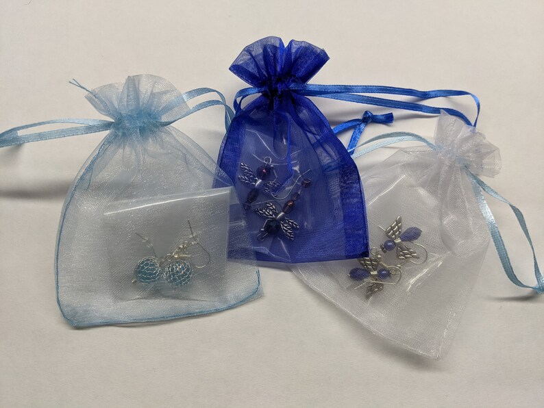 Puede incluir: Tres peque&ntilde;as bolsas de organza con cintas de diferentes colores. Cada bolsa contiene un par de pendientes. Una bolsa es azul, otra es blanca y la &uacute;ltima es azul claro.