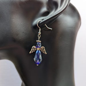 Puede incluir: Un par de pendientes colgantes con un dije de &aacute;ngel plateado y un colgante de l&aacute;grima de cristal azul oscuro.