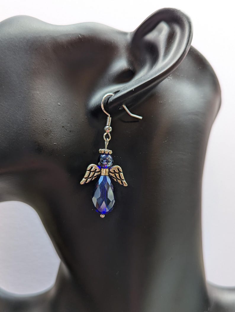 Puede incluir: Un par de pendientes colgantes con un dije de &aacute;ngel plateado y un colgante de l&aacute;grima de cristal azul oscuro.