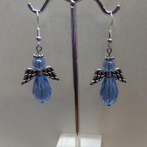 Puede incluir: Un par de pendientes colgantes con una l&aacute;grima de cristal azul claro y alas de &aacute;ngel plateadas.