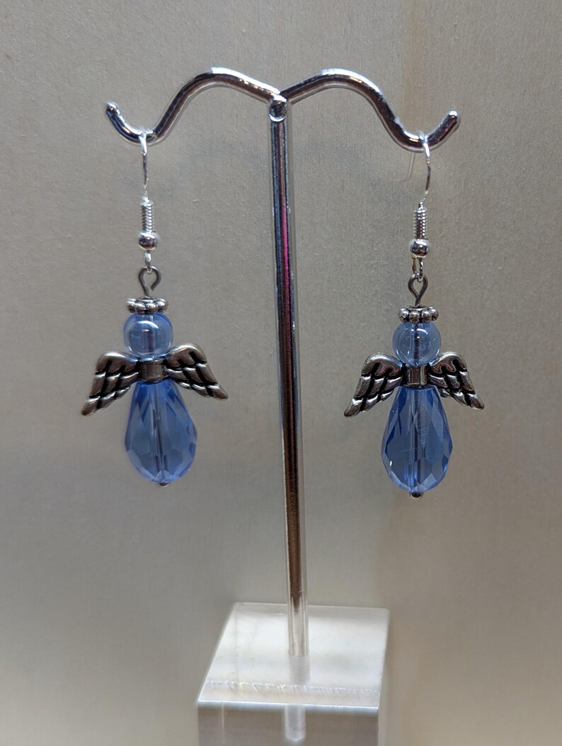 Puede incluir: Un par de pendientes colgantes con una l&aacute;grima de cristal azul claro y alas de &aacute;ngel plateadas.