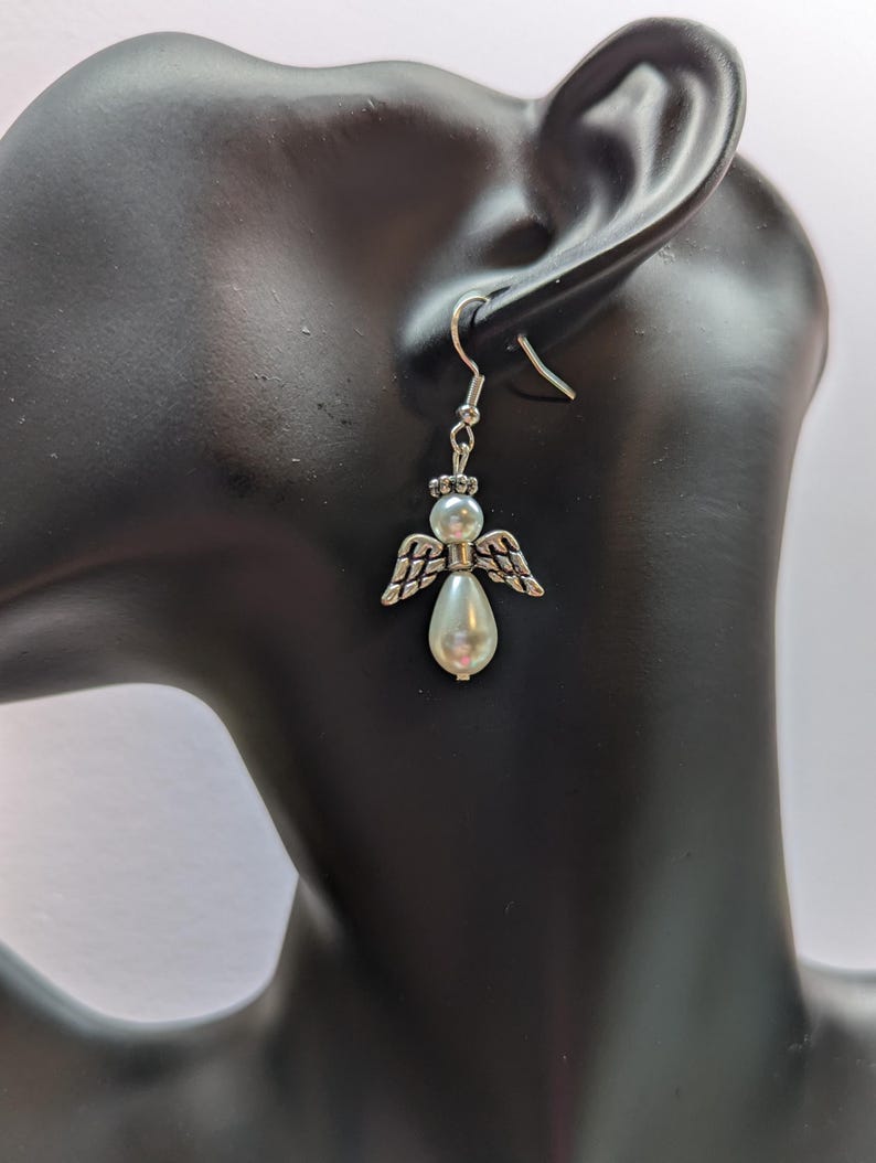 Puede incluir: Pendientes de plata con forma de &aacute;ngel y una perla blanca. Los pendientes est&aacute;n en la oreja de un maniqu&iacute;.