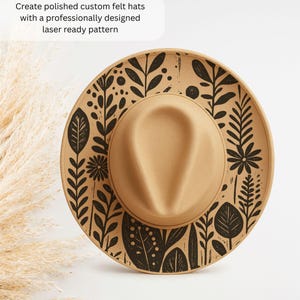 Op de afbeelding: Een beige vilten hoed met een zwart bloemenpatroon rond de rand. De hoed heeft een ronde kroon en een brede rand. Tekst op de afbeelding luidt: Maak gepolijste op maat gemaakte vilten hoeden met een professioneel ontworpen laser-klaar patroon.