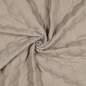 Op de afbeelding: Close-up van een beige gewatteerde stof met een herhalend patroon van verbonden, afgeronde vormen. De stof is zacht en heeft een lichte glans, met zichtbare stiksels die elke vorm omlijnen. De stof is in een spiraalvorm verzameld.