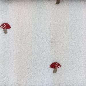 Könnte beinhalten: Ein weißer Sherpa-Fleece-Stoff mit einem wiederkehrenden Muster aus kleinen gestickten Pilzen. Jeder Pilz hat einen roten Hut mit weißen Punkten und einen hellbraunen Stiel. Die Textur des Stoffes ist dick, weich und lockig, was für ein flauschiges Aussehen sorgt.