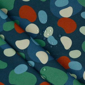 Op de afbeelding: Een close-up van een blauwe stof met een patroon van verschillende, abstracte vormen in tinten blauw, groen, crème en oranje. Waterdruppels zijn zichtbaar op het oppervlak van de stof.