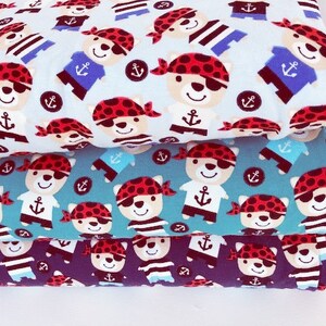 May include: Three stacks of fabric featuring a repeating pattern of cartoon pirate bears. The bears wear red bandanas and eye patches, with anchor and striped shirt details. The fabric colors are light blue, teal, and navy.
