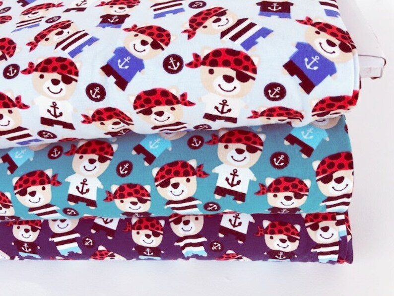 May include: Three stacks of fabric featuring a repeating pattern of cartoon pirate bears. The bears wear red bandanas and eye patches, with anchor and striped shirt details. The fabric colors are light blue, teal, and navy.