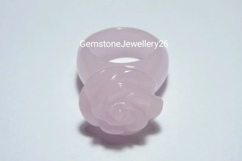 May include: A pale pink rose ring made of carved gemstone. The ring is a simple band with a rose carved on the top.