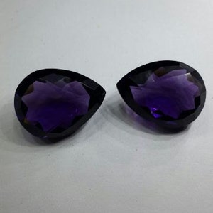 Pode incluir: Duas pedras preciosas facetadas em forma de p&ecirc;ra de cor p&uacute;rpura com um design de estrela.