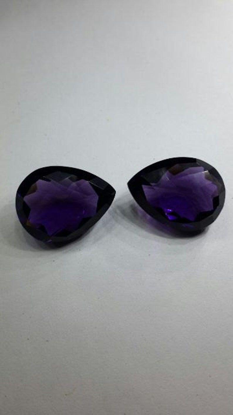Pode incluir: Duas pedras preciosas facetadas em forma de p&ecirc;ra de cor p&uacute;rpura com um design de estrela.