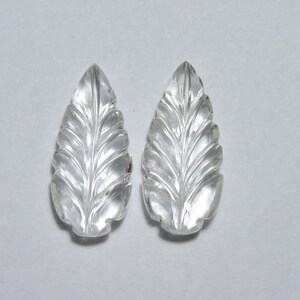 May include: Two clear quartz leaf-shaped pendants with intricate carvings. The pendants are smooth and polished, with a translucent appearance.