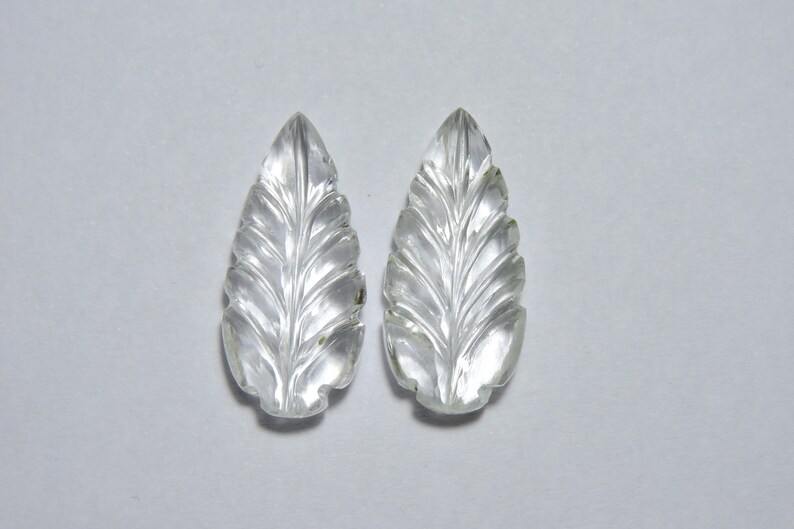 May include: Two clear quartz leaf-shaped pendants with intricate carvings. The pendants are smooth and polished, with a translucent appearance.