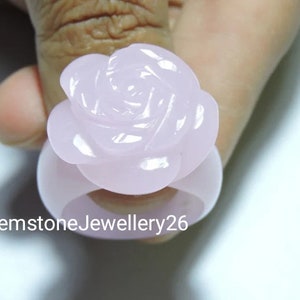 May include: A pale pink rose ring made of carved gemstone. The ring is worn on a finger.