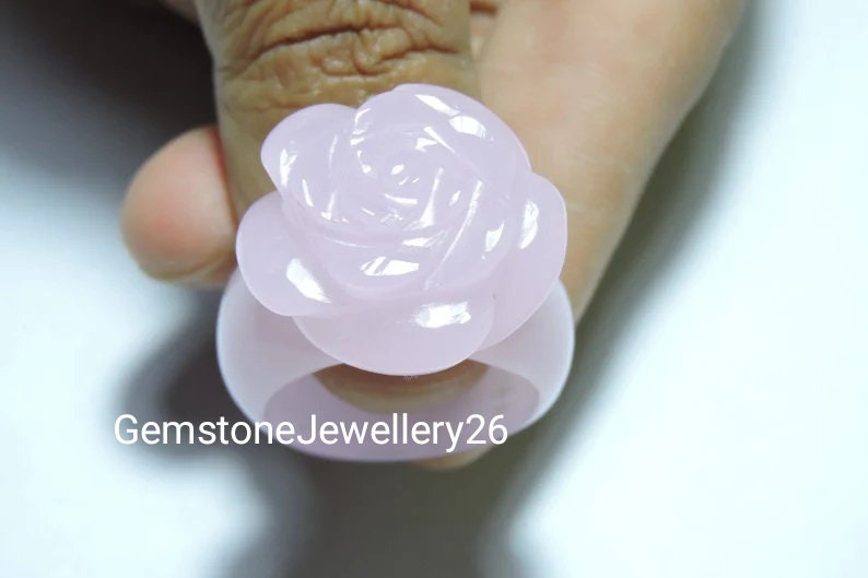 May include: A pale pink rose ring made of carved gemstone. The ring is worn on a finger.