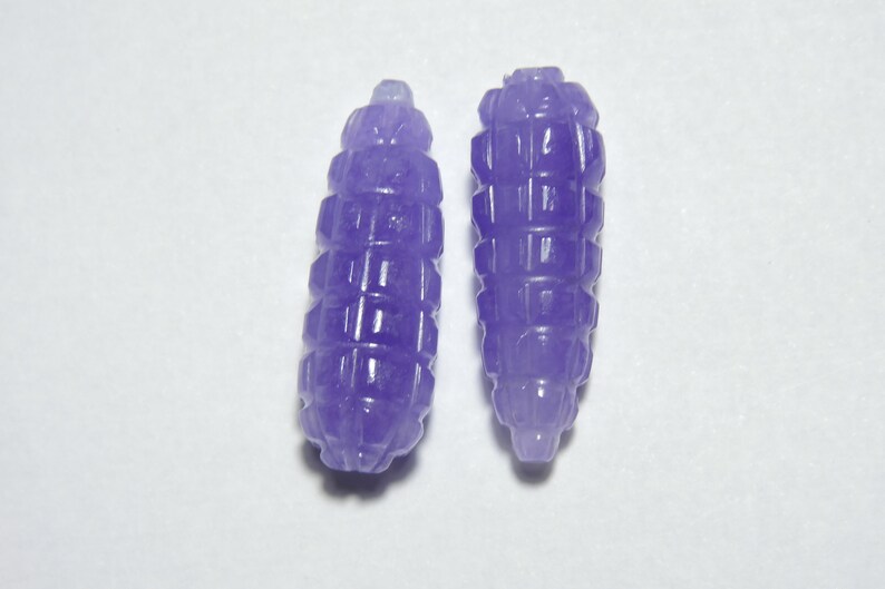 May include: Two purple jade beads with a textured, carved design. The beads are shaped like small, elongated barrels.