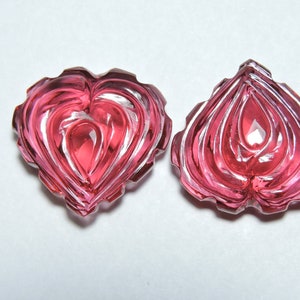 May include: Two pink heart-shaped carved gemstones with intricate designs. The gemstones are translucent and have a glossy finish.