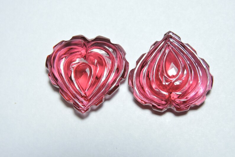May include: Two pink heart-shaped carved gemstones with intricate designs. The gemstones are translucent and have a glossy finish.