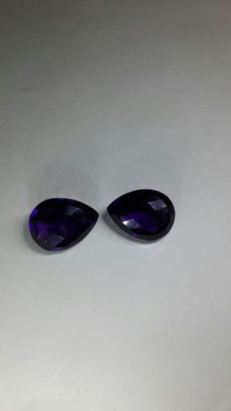 Pode incluir: Duas pedras preciosas de ametista facetadas em forma de l&aacute;grima, cada uma com um tom roxo profundo.