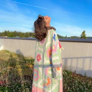 Pode incluir: Uma pessoa vestindo uma colcha vintage como um vestido, em pé em um campo de plantas verdes com flores brancas. A colcha é feita de tecido patchwork com um padrão floral em tons de rosa, verde e laranja.