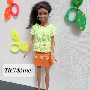 Peut inclure: Une poupée vêtue d'un haut en tricot vert citron et d'une jupe orange avec des motifs de cœurs verts. La poupée porte des chaussures jaunes et a les cheveux noirs. Des élastiques à cheveux avec des motifs de feuilles sont également visibles.