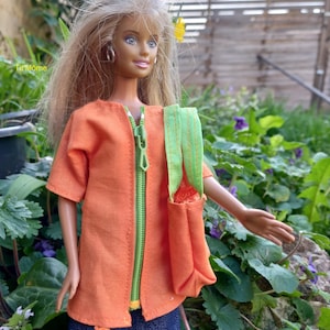 Peut inclure: Une poupée vêtue d'une veste orange avec une fermeture éclair verte et un sac assorti à bretelles vertes. La poupée porte également un jean bleu avec des motifs de fleurs colorées. La poupée a les cheveux blonds et se trouve dans un jardin.