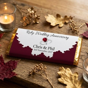 May include: A chocolate bar with a white and claret label, celebrating a Ruby Wedding Anniversary. The label features a ring design and the names "Chris & Phil" with the date "3rd November 2027". The bar is surrounded by candles and decorative leaves.