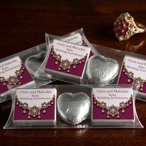 May include: A selection of chocolates in transparent packaging, featuring heart-shaped sweets and rectangular chocolates. The rectangular chocolates have a burgundy label with gold and white decorative elements and the text "Chris and Malcolm Ruby Wedding Anniversary". A gold ring with red stones is in the background.
