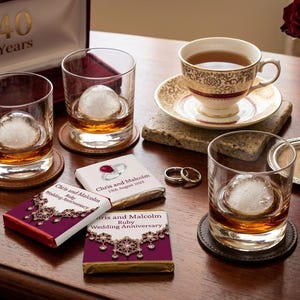 May include: A collection of items celebrating a ruby wedding anniversary. The image includes whiskey glasses with ice spheres, a teacup, chocolates, and wedding rings. A red rose in a vase and a box with "40 Years" are also visible.