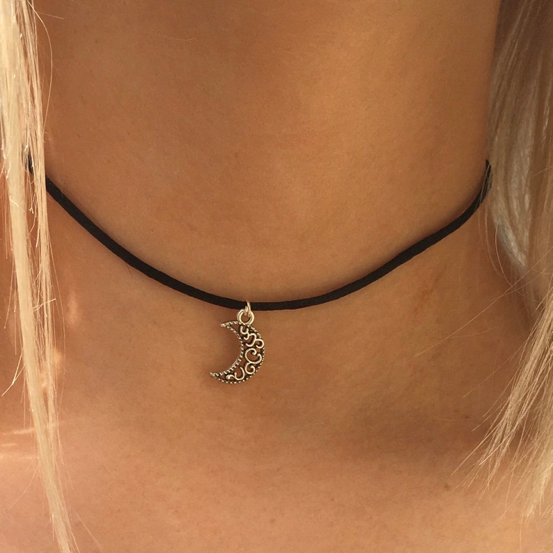 Puede incluir: Un collar gargantilla negro con un colgante de luna creciente plateado. El colgante tiene un dise&ntilde;o delicado e intrincado.