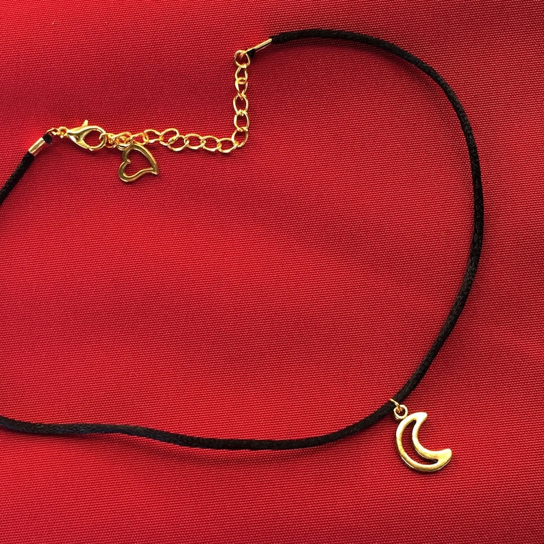 Puede incluir: Un collar gargantilla negro con un dije de media luna dorada y un dije de coraz&oacute;n dorado en el cierre.