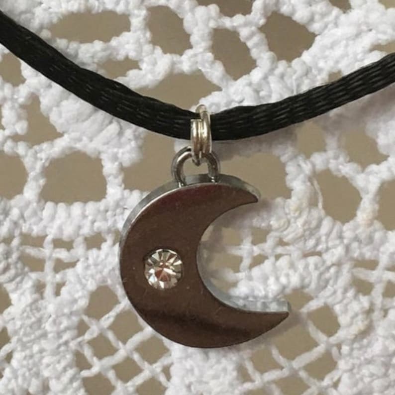 Puede incluir: Un collar con un colgante de media luna de plata con un strass transparente en un cord&oacute;n negro.