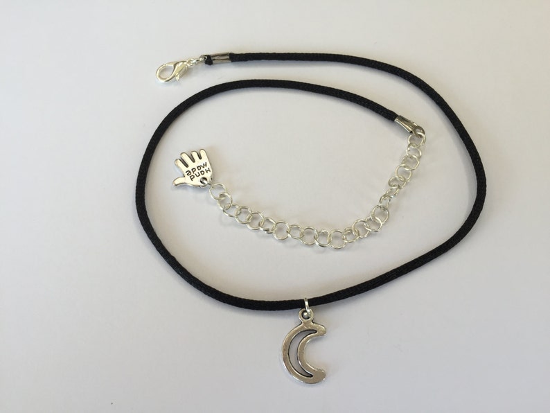 Puede incluir: Collar gargantilla negro con un dije de plata en forma de media luna y un dije de plata en forma de mano con las palabras "Happy Buddha" inscritas en &eacute;l.