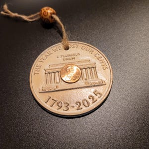 May include: A round, beige-colored pendant with the text "THE YEAR WE LOST OUR CENTS" and "1793-2025" is displayed. A copper penny rests on the pendant. A wooden bead is attached to the pendant with a twine string.