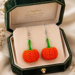 May include: A pair of orange crochet pumpkin earrings with green stems and silver-toned metal hooks, presented inside a dark green velvet jewelry box with the text HANDMADE WITH LOVE printed in gold on the interior lid lining.