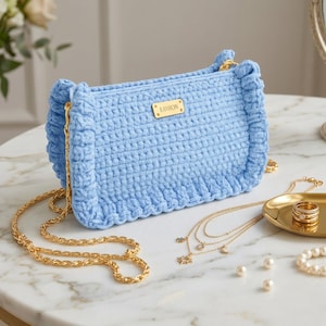 Könnte beinhalten: Eine hellblaue gehäkelte Handtasche in rechteckiger Form mit gerüschten Seitenkanten und einem goldfarbenen Metallkettenriemen. Ein kleines rechteckiges goldfarbenes Metallschild mit der Aufschrift EAVHON ist mittig auf der Vorderseite angebracht.